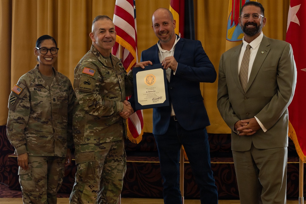 A military award or recognition ceremony is taking place in a formal indoor setting. A uniformed U.S. Army officer is shaking hands with a civilian man in a blue suit who is holding a certificate or commendation document. Two additional individuals — a female soldier and a civilian man in a tan suit — stand alongside as witnesses.