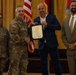 A military award or recognition ceremony is taking place in a formal indoor setting. A uniformed U.S. Army officer is shaking hands with a civilian man in a blue suit who is holding a certificate or commendation document. Two additional individuals — a female soldier and a civilian man in a tan suit — stand alongside as witnesses.