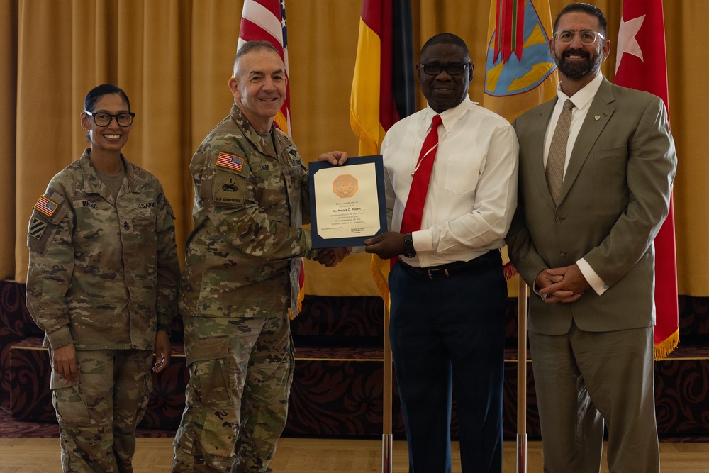 A formal military award ceremony is taking place in what appears to be a ceremonial hall. A U.S. Army soldier in uniform is presenting a framed certificate or award to a civilian man in a white shirt and red tie. Two additional individuals — a female soldier and a civilian man in a suit — stand alongside as witnesses.