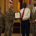 A formal military award ceremony is taking place in what appears to be a ceremonial hall. A U.S. Army soldier in uniform is presenting a framed certificate or award to a civilian man in a white shirt and red tie. Two additional individuals — a female soldier and a civilian man in a suit — stand alongside as witnesses.