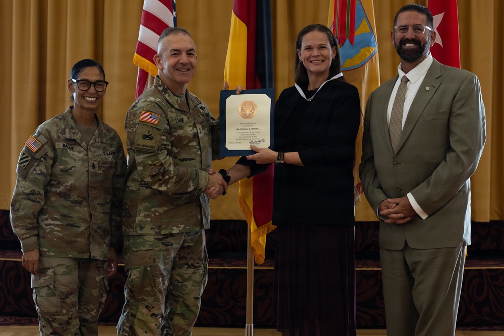 A formal military recognition ceremony is taking place in what appears to be a ceremonial hall. A U.S. Army officer is presenting a certificate to a civilian woman, with two other individuals — a female soldier and a civilian man — standing alongside them. Multiple flags, including the U.S. flag and what appear to be state and unit flags, are visible in the background.