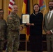 A formal military recognition ceremony is taking place in what appears to be a ceremonial hall. A U.S. Army officer is presenting a certificate to a civilian woman, with two other individuals — a female soldier and a civilian man — standing alongside them. Multiple flags, including the U.S. flag and what appear to be state and unit flags, are visible in the background.