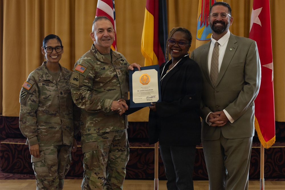 A formal military award ceremony is taking place, with a uniformed Army officer presenting a framed certificate to a civilian woman. Four individuals are posed together: two in U.S. Army combat uniforms, one civilian woman receiving the award, and one civilian man in a suit. The setting appears to be an official venue with U.S., German, and military unit flags displayed in the background.