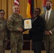 A formal military award ceremony is taking place, with a uniformed Army officer presenting a framed certificate to a civilian woman. Four individuals are posed together: two in U.S. Army combat uniforms, one civilian woman receiving the award, and one civilian man in a suit. The setting appears to be an official venue with U.S., German, and military unit flags displayed in the background.