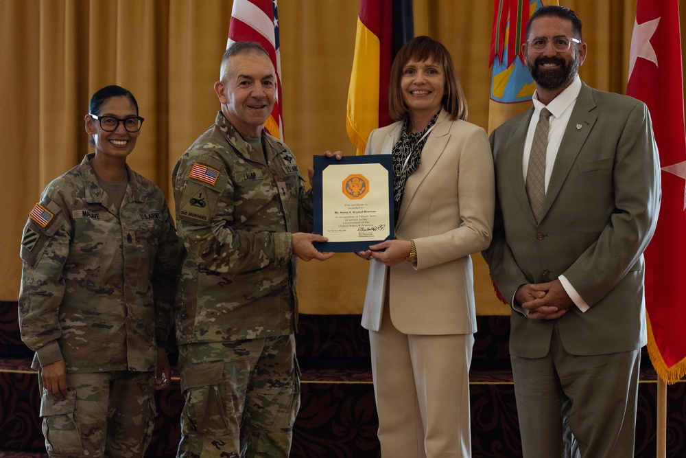 A formal military ceremony in which a U.S. Army general officer presents a certificate or award to a civilian woman in a beige suit. Four individuals are posed together in front of flags including the U.S. flag and what appears to be unit/organizational flags. The setting appears to be an official military function room with gold drapery in the background.