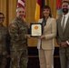 A formal military ceremony in which a U.S. Army general officer presents a certificate or award to a civilian woman in a beige suit. Four individuals are posed together in front of flags including the U.S. flag and what appears to be unit/organizational flags. The setting appears to be an official military function room with gold drapery in the background.