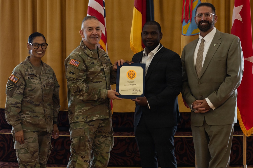 A formal military recognition ceremony is taking place, with a U.S. Army officer presenting a certificate or commendation to a civilian man in a dark suit. Four individuals are posed together: two in U.S. Army combat uniforms, one civilian recipient, and one civilian in a tan suit. The setting appears to be an official military venue with multiple flags including the U.S. flag and what appears to be a German flag in the background.