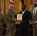 A formal military recognition ceremony is taking place, with a U.S. Army officer presenting a certificate or commendation to a civilian man in a dark suit. Four individuals are posed together: two in U.S. Army combat uniforms, one civilian recipient, and one civilian in a tan suit. The setting appears to be an official military venue with multiple flags including the U.S. flag and what appears to be a German flag in the background.