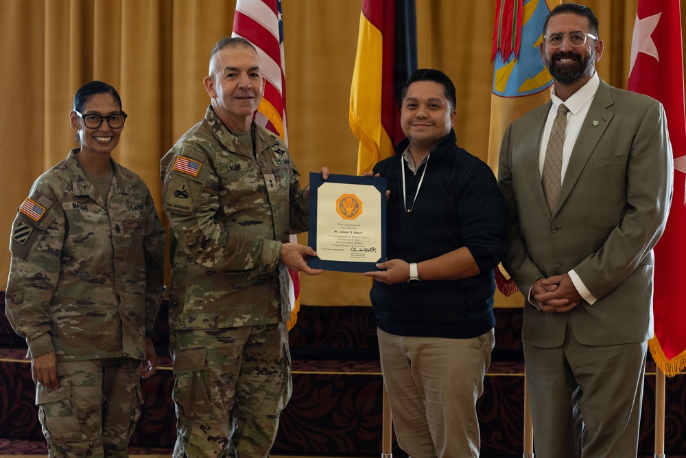 A formal military award ceremony is taking place, with a U.S. Army officer presenting a certificate to a civilian recipient. Two military personnel in Army Combat Uniforms (ACU) and one civilian in a suit are also present. The setting appears to be an official venue with multiple flags displayed in the background.