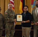 A formal military award ceremony is taking place, with a U.S. Army officer presenting a certificate to a civilian recipient. Two military personnel in Army Combat Uniforms (ACU) and one civilian in a suit are also present. The setting appears to be an official venue with multiple flags displayed in the background.