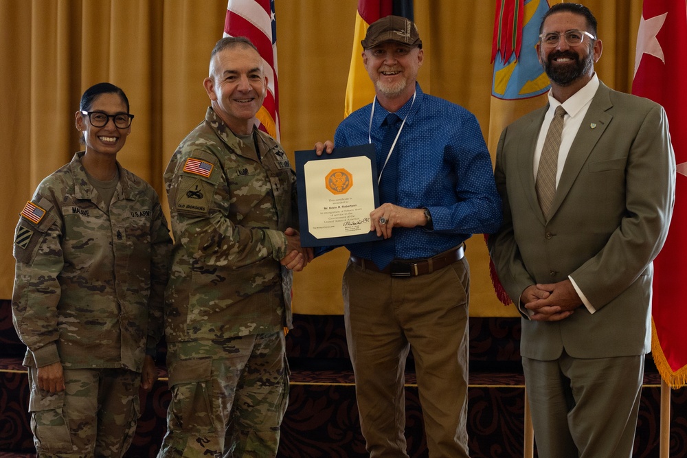 A formal military recognition ceremony in which a uniformed U.S. Army officer is presenting a certificate to a civilian male. Four individuals are posed together for a photo: two in U.S. Army combat uniforms, one civilian recipient holding the certificate, and one civilian in a suit. The setting includes U.S. and military unit flags in the background against gold/yellow draping.