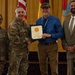 A formal military recognition ceremony in which a uniformed U.S. Army officer is presenting a certificate to a civilian male. Four individuals are posed together for a photo: two in U.S. Army combat uniforms, one civilian recipient holding the certificate, and one civilian in a suit. The setting includes U.S. and military unit flags in the background against gold/yellow draping.