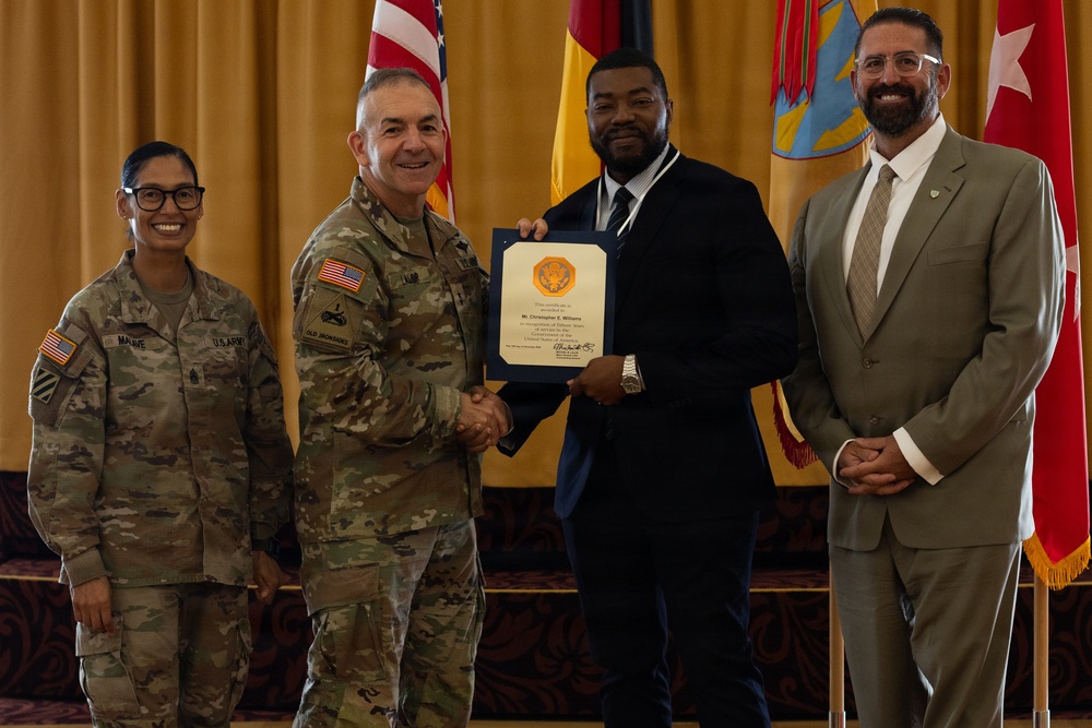 A formal military award ceremony showing four individuals posing for a photo. A U.S. Army officer in camouflage uniform is shaking hands with a civilian man in a dark suit while both hold a certificate or award document. Two other individuals — a female soldier and a civilian man in a tan suit — stand on either side.