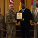 A formal military award ceremony showing four individuals posing for a photo. A U.S. Army officer in camouflage uniform is shaking hands with a civilian man in a dark suit while both hold a certificate or award document. Two other individuals — a female soldier and a civilian man in a tan suit — stand on either side.