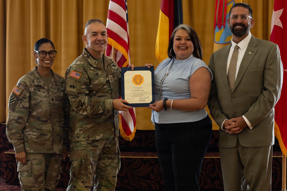 A formal military recognition ceremony is taking place, with a U.S. Army officer presenting a certificate or award to a civilian woman. Four individuals are posed together in front of American and military unit flags in what appears to be an official ceremony hall. Two military personnel in Army Combat Uniforms (ACU) and two civilians are present.
