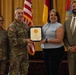 A formal military recognition ceremony is taking place, with a U.S. Army officer presenting a certificate or award to a civilian woman. Four individuals are posed together in front of American and military unit flags in what appears to be an official ceremony hall. Two military personnel in Army Combat Uniforms (ACU) and two civilians are present.