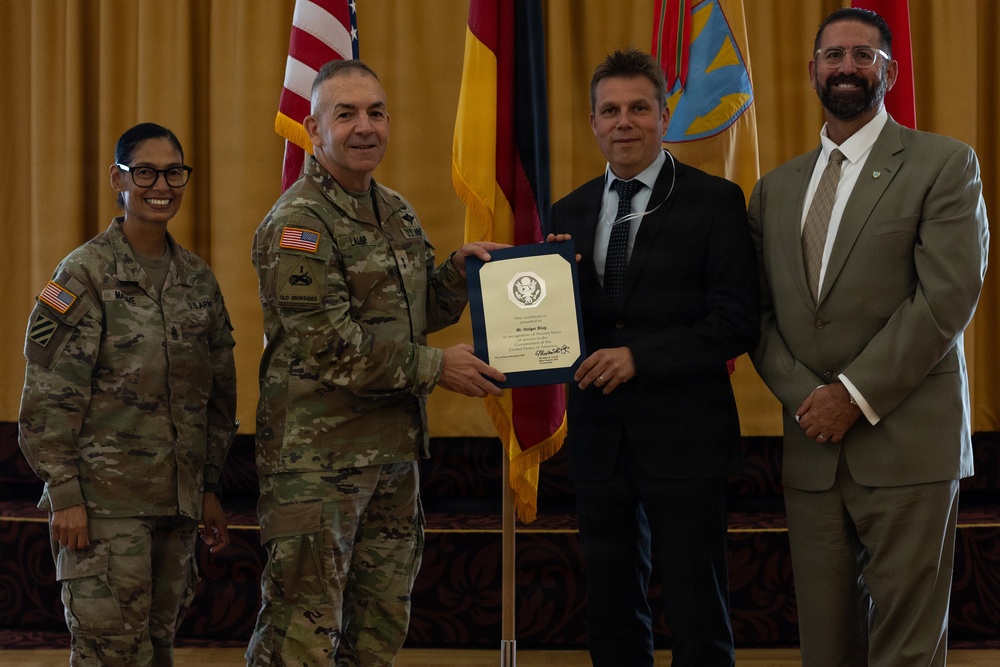 A formal military award ceremony is taking place, with a U.S. Army officer presenting a framed certificate to a civilian male. Four individuals are present: two in U.S. Army combat uniforms, one civilian male receiving the award, and one civilian male in a suit standing to the right. Flags including the U.S. flag and German flag are visible in the background, suggesting a U.S. military installation in Germany.