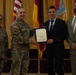 A formal military award ceremony is taking place, with a U.S. Army officer presenting a framed certificate to a civilian male. Four individuals are present: two in U.S. Army combat uniforms, one civilian male receiving the award, and one civilian male in a suit standing to the right. Flags including the U.S. flag and German flag are visible in the background, suggesting a U.S. military installation in Germany.