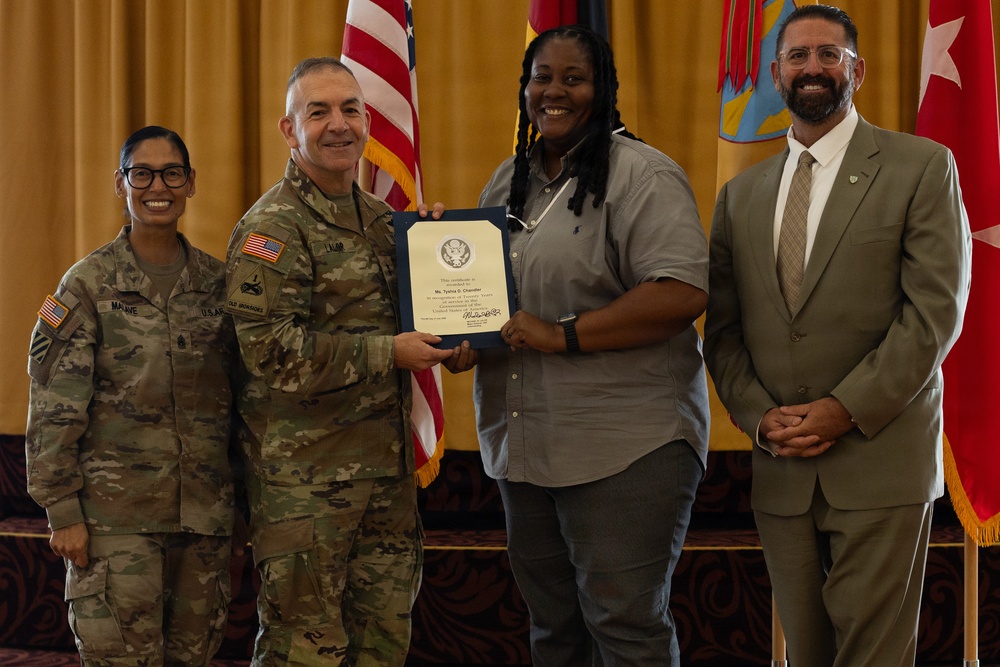 A formal military recognition ceremony is taking place indoors, where a uniformed Army officer is presenting a certificate or award to a civilian woman. Three other individuals are present, including a female soldier and a civilian man in a suit. The setting includes American and military flags, suggesting an official government or Army installation event.