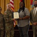 A formal military recognition ceremony is taking place indoors, where a uniformed Army officer is presenting a certificate or award to a civilian woman. Three other individuals are present, including a female soldier and a civilian man in a suit. The setting includes American and military flags, suggesting an official government or Army installation event.