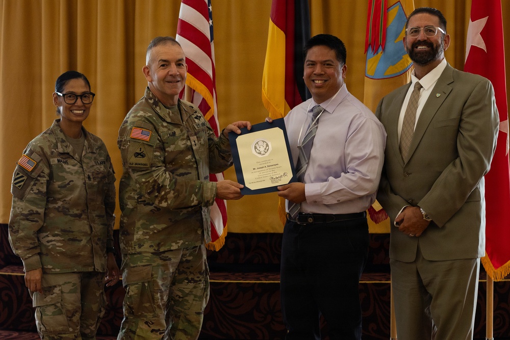 A military award or recognition ceremony is taking place, with a uniformed U.S. Army soldier presenting a framed certificate to a civilian man. Four individuals are present: two in U.S. Army combat uniforms and two in civilian attire. Multiple flags, including the U.S. flag and what appears to be a German flag, are visible in the background.