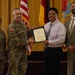 A military award or recognition ceremony is taking place, with a uniformed U.S. Army soldier presenting a framed certificate to a civilian man. Four individuals are present: two in U.S. Army combat uniforms and two in civilian attire. Multiple flags, including the U.S. flag and what appears to be a German flag, are visible in the background.