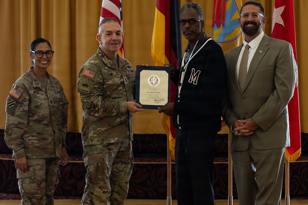 A formal military award ceremony is taking place indoors, where a uniformed Army soldier is presenting a framed certificate to a civilian man wearing a varsity-style jacket with the letter 'M'. Three other individuals are present, including a female soldier in uniform and a civilian man in a suit, standing before flags including the American flag and what appears to be a German flag.