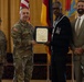 A formal military award ceremony is taking place indoors, where a uniformed Army soldier is presenting a framed certificate to a civilian man wearing a varsity-style jacket with the letter 'M'. Three other individuals are present, including a female soldier in uniform and a civilian man in a suit, standing before flags including the American flag and what appears to be a German flag.