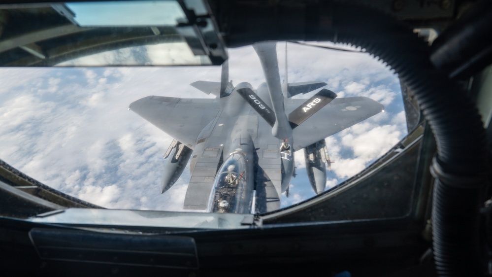 This image is taken from the boom operator's station of a tanker aircraft, showing an aerial refueling operation in progress. A fighter jet, identifiable as an F-15 Eagle, is positioned directly below the refueling boom, receiving fuel at altitude above a cloud layer. The markings 'ARS' and the number '909' are visible on the tanker's wing or fuselage.