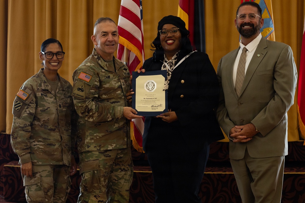 A formal military award ceremony is taking place in what appears to be an official venue. A civilian woman identified on the certificate as 'Ms. Rasheeda A. Hall' is receiving a certificate from a military officer in Army Combat Uniform, flanked by two other individuals. The American flag and additional military/organizational flags are visible in the background.