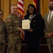 A formal military award ceremony is taking place in what appears to be an official venue. A civilian woman identified on the certificate as 'Ms. Rasheeda A. Hall' is receiving a certificate from a military officer in Army Combat Uniform, flanked by two other individuals. The American flag and additional military/organizational flags are visible in the background.
