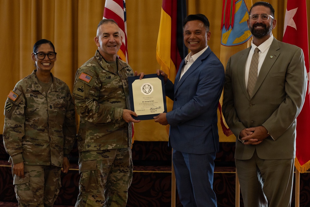 A formal military award ceremony is taking place, with a U.S. Army officer presenting a framed certificate to a civilian man in a blue suit. Four individuals are present: two in U.S. Army combat uniforms, one civilian recipient, and one civilian observer in a tan suit. Multiple flags including the U.S. flag and what appears to be a New Mexico state flag are visible in the background.