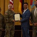 A formal military award ceremony is taking place, with a U.S. Army officer presenting a framed certificate to a civilian man in a blue suit. Four individuals are present: two in U.S. Army combat uniforms, one civilian recipient, and one civilian observer in a tan suit. Multiple flags including the U.S. flag and what appears to be a New Mexico state flag are visible in the background.