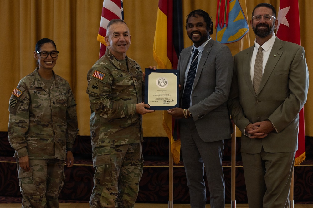 A formal military award ceremony is taking place in what appears to be an official venue. A U.S. Army officer in camouflage uniform is presenting a framed certificate to a civilian man in a gray suit, while two other individuals stand alongside them. Multiple flags, including the U.S. flag and military unit flags, are visible in the background.