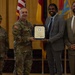 A formal military award ceremony is taking place in what appears to be an official venue. A U.S. Army officer in camouflage uniform is presenting a framed certificate to a civilian man in a gray suit, while two other individuals stand alongside them. Multiple flags, including the U.S. flag and military unit flags, are visible in the background.