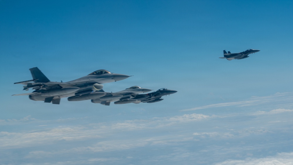 The image shows a formation of military fighter jets flying in clear blue sky at high altitude. Multiple aircraft are visible in close formation on the left side, with one aircraft flying separately to the upper right. Cloud cover is visible below the aircraft, suggesting high-altitude flight.