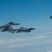 The image shows a formation of military fighter jets flying in clear blue sky at high altitude. Multiple aircraft are visible in close formation on the left side, with one aircraft flying separately to the upper right. Cloud cover is visible below the aircraft, suggesting high-altitude flight.