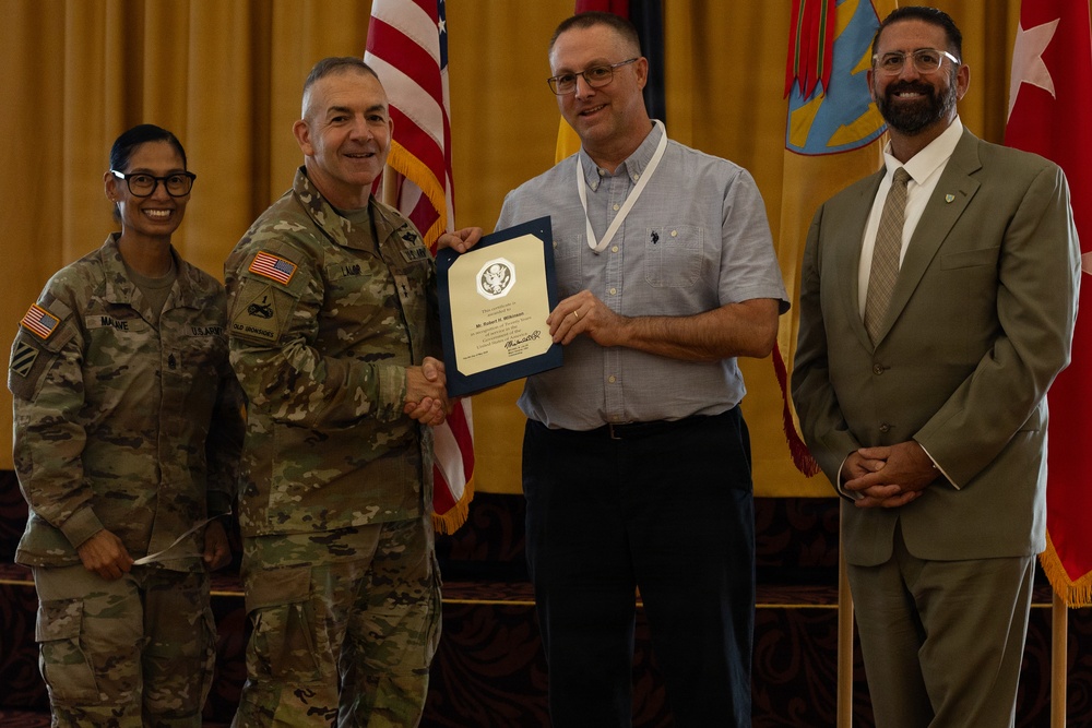 A military award ceremony is taking place in a formal indoor setting. A U.S. Army officer in camouflage uniform is presenting a certificate or commendation to a civilian male while shaking hands. Two other individuals — a female soldier and a civilian male in a suit — stand alongside as witnesses.