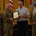 A military award ceremony is taking place in a formal indoor setting. A U.S. Army officer in camouflage uniform is presenting a certificate or commendation to a civilian male while shaking hands. Two other individuals — a female soldier and a civilian male in a suit — stand alongside as witnesses.