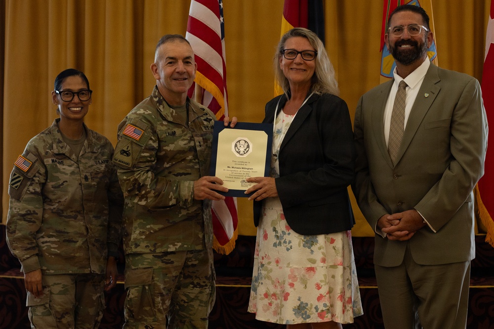 A formal military ceremony in which a U.S. Army officer is presenting a certificate or award to a civilian woman. Four individuals are posed together for a photo: two in U.S. Army combat uniforms and two in civilian attire. The setting appears to be an official venue with American and military flags in the background.