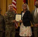 A formal military ceremony in which a U.S. Army officer is presenting a certificate or award to a civilian woman. Four individuals are posed together for a photo: two in U.S. Army combat uniforms and two in civilian attire. The setting appears to be an official venue with American and military flags in the background.