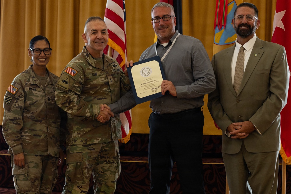 A formal military award ceremony is taking place, with a U.S. Army officer presenting a certificate or commendation to a civilian recipient. Four individuals are present: two in U.S. Army combat uniforms, one civilian in a gray shirt receiving the award, and another civilian in a tan suit standing to the right. The setting includes U.S. and military unit flags in the background, suggesting an official installation or headquarters venue.