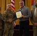 A formal military award ceremony is taking place, with a U.S. Army officer presenting a certificate or commendation to a civilian recipient. Four individuals are present: two in U.S. Army combat uniforms, one civilian in a gray shirt receiving the award, and another civilian in a tan suit standing to the right. The setting includes U.S. and military unit flags in the background, suggesting an official installation or headquarters venue.