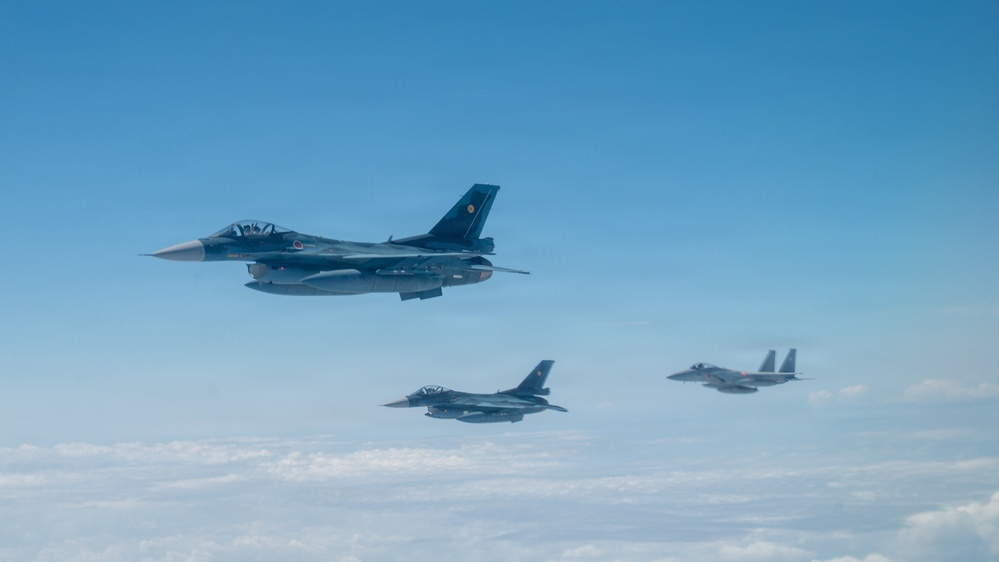 Three military jet fighters are flying in a loose formation at altitude above a cloud layer against a clear blue sky. The formation consists of two single-engine fighters that appear to be F-2 or F-16 variants in the foreground and middle, and a twin-engine fighter that appears to be an F-15 variant on the right. The aircraft display markings consistent with the Japan Air Self-Defense Force (JASDF).