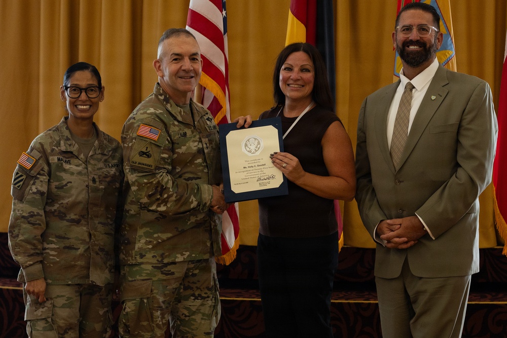 A formal military ceremony in which a U.S. Army officer presents a certificate or award to a civilian woman. Four individuals are posed together in front of American and military unit flags against a gold curtain backdrop. The occasion appears to be a recognition or commendation event.