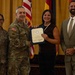 A formal military ceremony in which a U.S. Army officer presents a certificate or award to a civilian woman. Four individuals are posed together in front of American and military unit flags against a gold curtain backdrop. The occasion appears to be a recognition or commendation event.