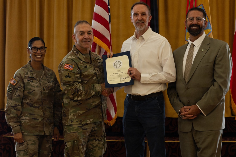 A formal military ceremony in which a U.S. Army officer is presenting a certificate or award to a civilian male. Four individuals are present: two uniformed Army soldiers, one civilian recipient, and one civilian observer in a suit. The setting appears to be an official venue with American and military flags in the background.