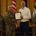 A formal military ceremony in which a U.S. Army officer is presenting a certificate or award to a civilian male. Four individuals are present: two uniformed Army soldiers, one civilian recipient, and one civilian observer in a suit. The setting appears to be an official venue with American and military flags in the background.