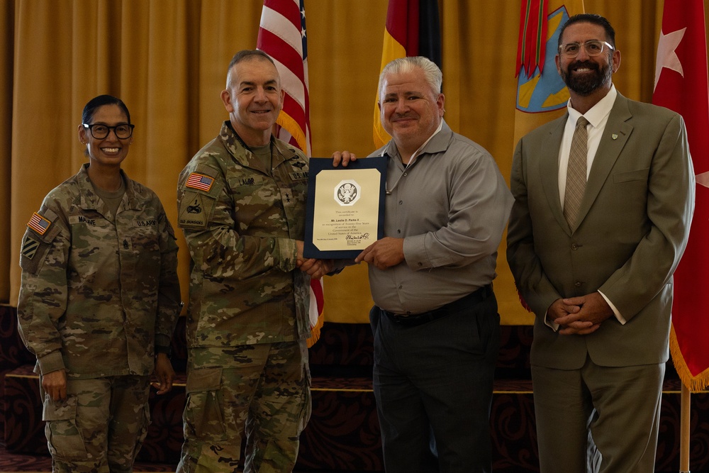 A military award or recognition ceremony is taking place indoors, with a uniformed U.S. Army officer presenting a framed certificate or commendation to a civilian male. Four individuals are posed together for a photo, including two military personnel in Army Combat Uniforms and two civilians. The setting appears to be a formal military venue with U.S. and unit flags displayed in the background.