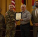 A military award or recognition ceremony is taking place indoors, with a uniformed U.S. Army officer presenting a framed certificate or commendation to a civilian male. Four individuals are posed together for a photo, including two military personnel in Army Combat Uniforms and two civilians. The setting appears to be a formal military venue with U.S. and unit flags displayed in the background.